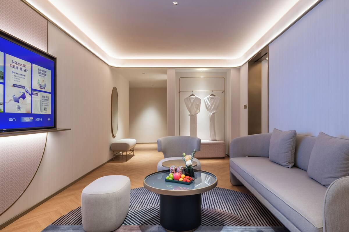 Ett modernt hotellrum med en platt-TV som är monterad på väggen, ett runt soffbord med frukt och en vas, en grå soffa, en grå fåtölj och en liten pall.