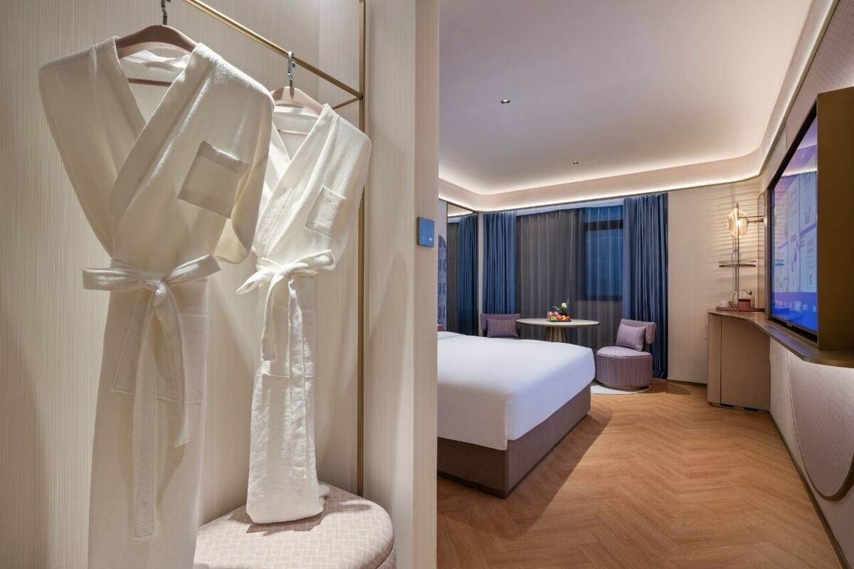 Ett hotellrum med en stor säng, ett litet bord med en stol, en tv och en klädställning med badrockar.