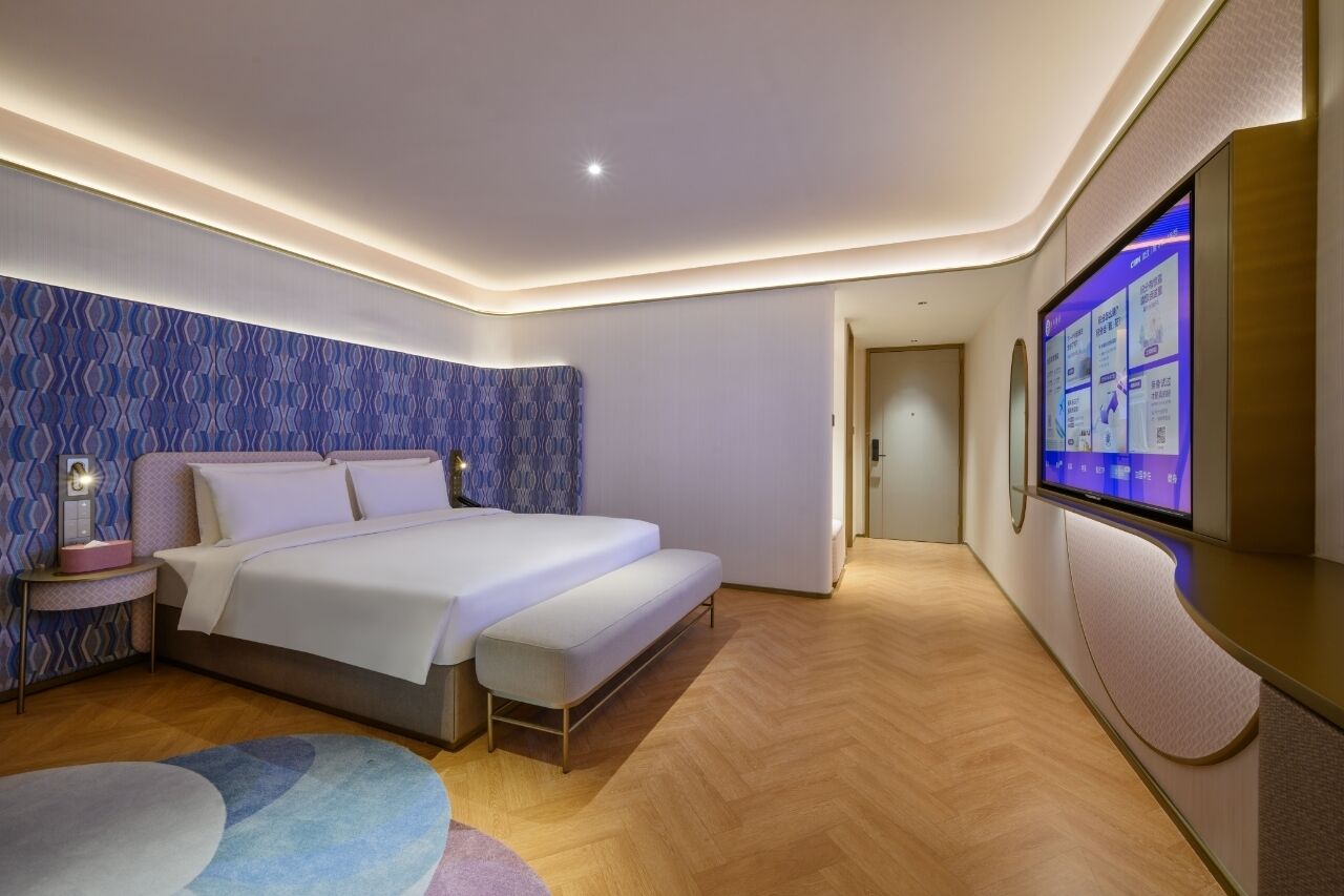 A modern hotel room with a large bed, a bedside table with a lamp, a flat-screen TV mounted on the wall, and a wooden floor.