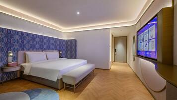A modern hotel room with a large bed, a bedside table with a lamp, a flat-screen TV mounted on the wall, and a wooden floor.