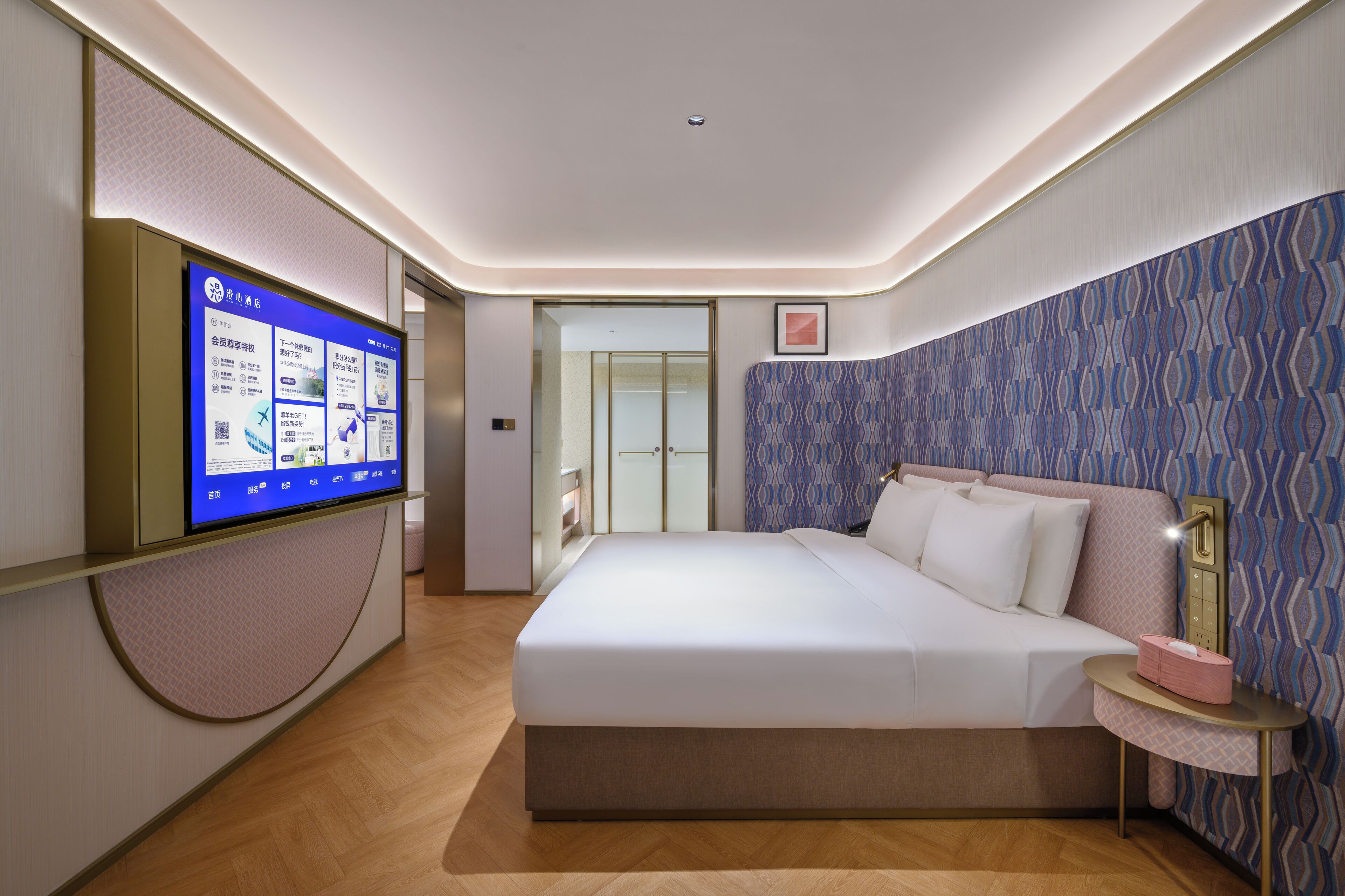 A modern hotel room with a large bed, a bedside table with a lamp, a flat-screen TV mounted on the wall, and a patterned wallpaper.