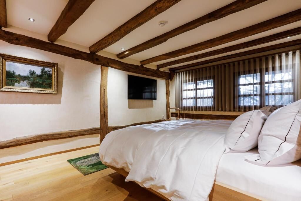 Een slaapkamer met zichtbare houten balken, een bed met witte beddengoed, een ingelijste foto aan de muur en een raam met jaloezieën.
