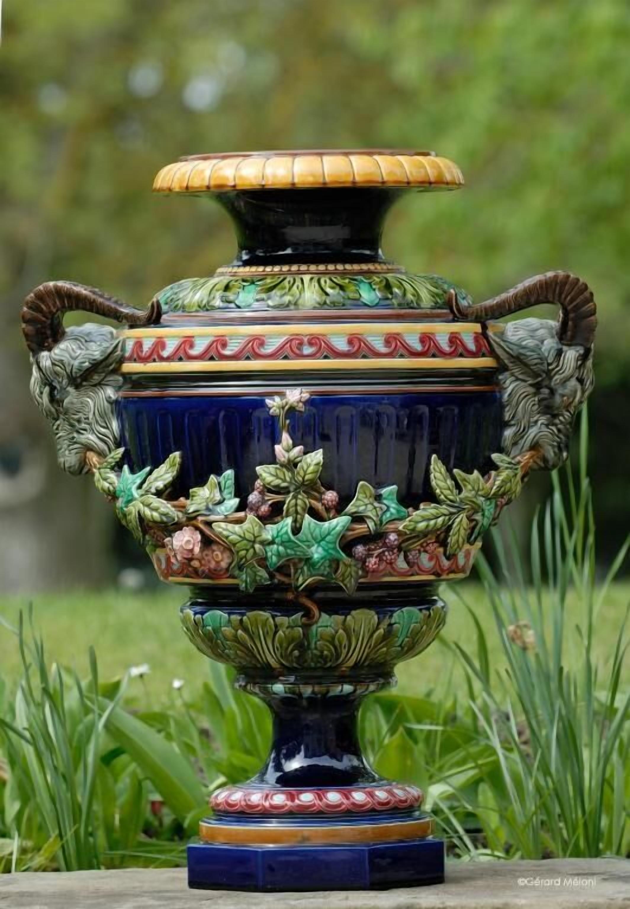 A decorative ceramic urn with intricate floral and leaf patterns, set on a stone base.
