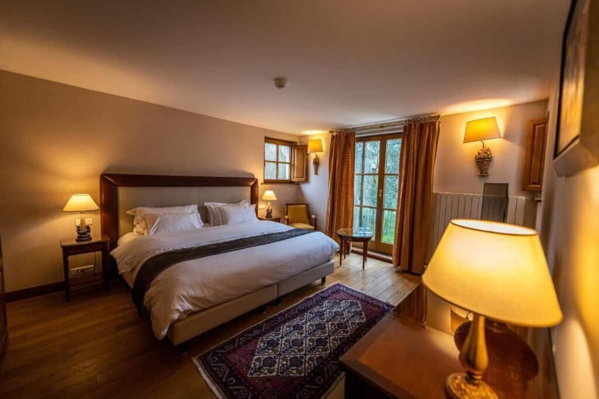 Une chambre d’hôtel avec un grand lit, deux tables de chevet avec des lampes, une petite table avec une chaise et une fenêtre avec des rideaux.