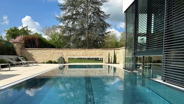 A swimming pool with a stone wall and a modern building with horizontal blinds. - Auberge Saint Walfrid  (Sarreguemines)