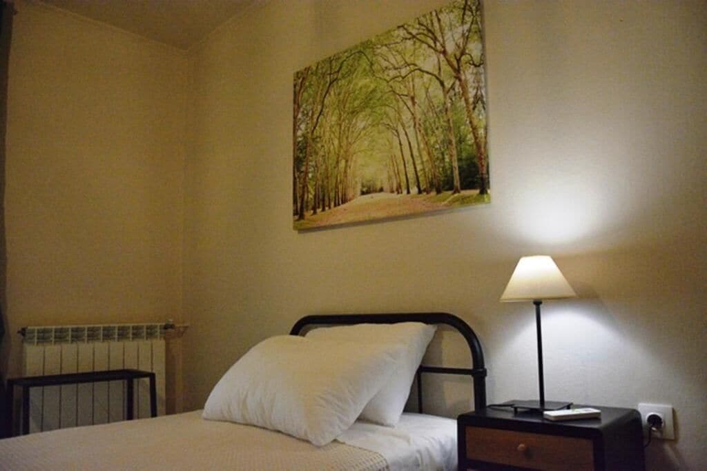 Ein Schlafzimmer mit einem Bett, einem Nachttisch mit Lampe und einem Waldgemälde an der Wand.