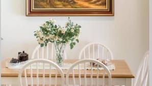 Un comedor con una mesa de madera, sillas blancas, un jarrón de flores y un cuadro de paisaje en la pared.
