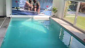 Ein Swimmingpool mit einem Wandgemälde von zwei Personen in einem Boot, einer Topfpflanze und Blick auf einen Garten durch eine Glastür.