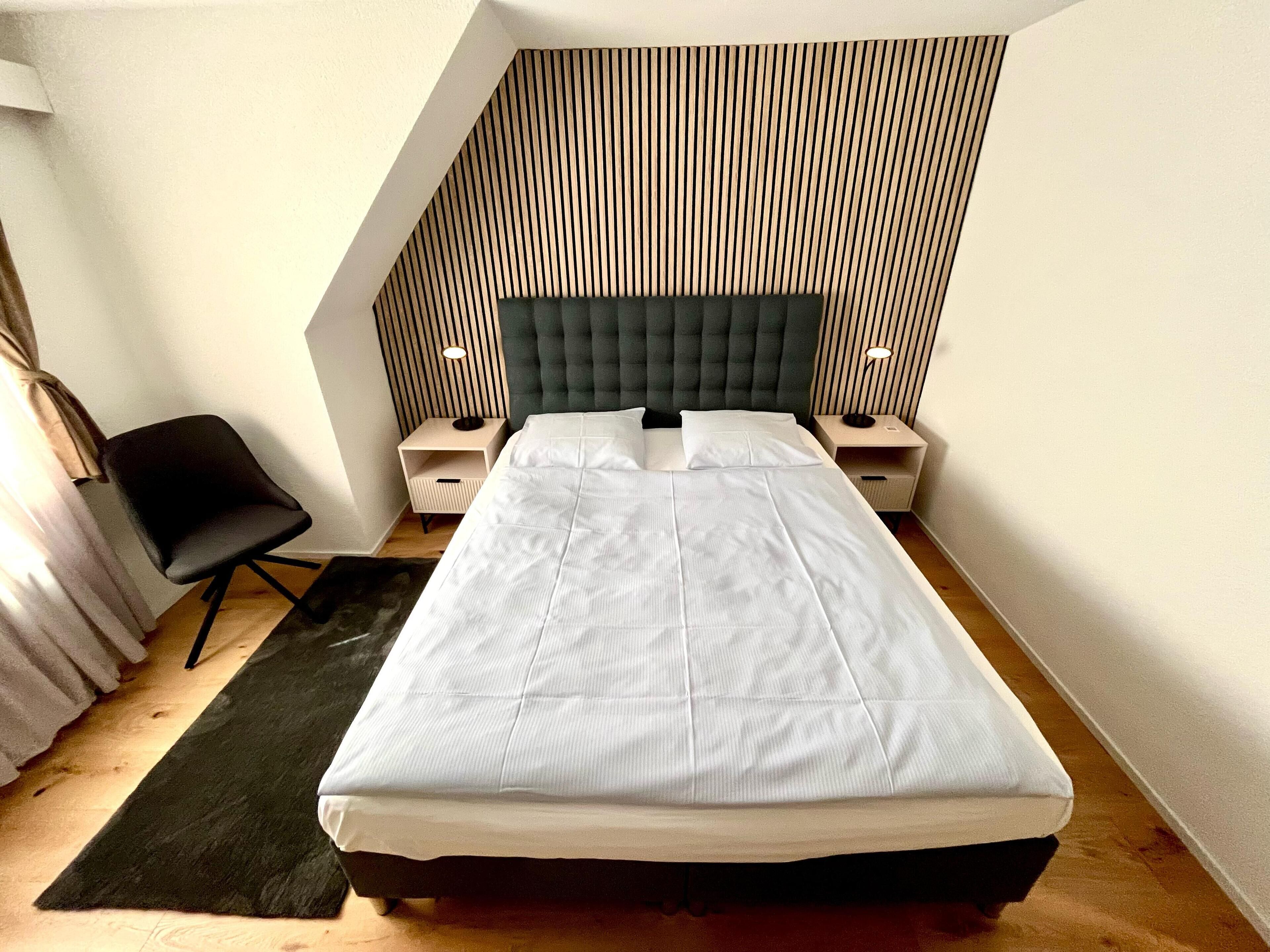 A modern bedroom with a large bed, two bedside tables, a chair, and a sloped ceiling with wooden paneling.