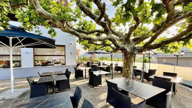 Una terrazza esterna con tavoli e sedie, circondata da alberi e un edificio con finestre.