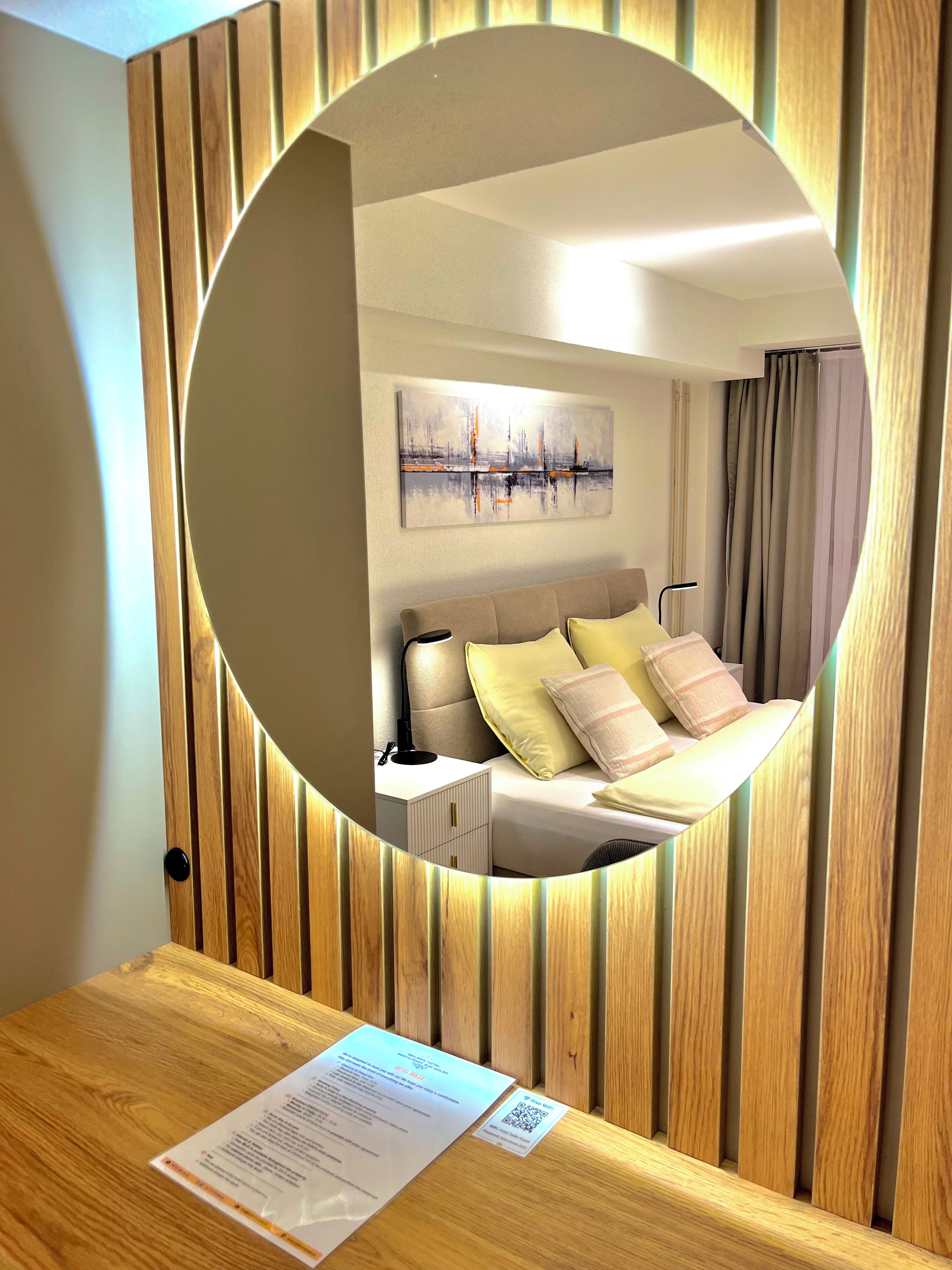 Ein Hotelzimmer mit einer Wandpaneele aus Holz, einem runden Spiegel, der den Raum reflektiert, einem Bett mit Kissen, einem Nachttisch und einem Bild an der Wand.