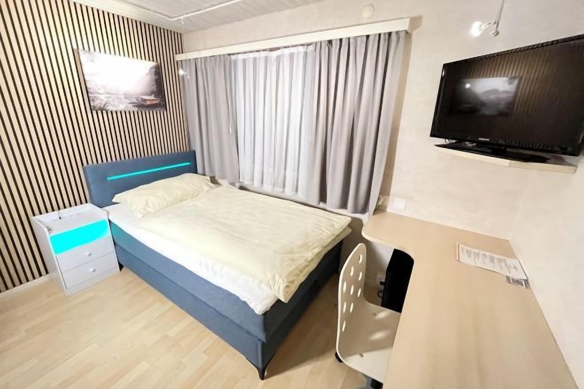 Una habitación de hotel con cama, escritorio, silla, televisión y ventana con cortinas.