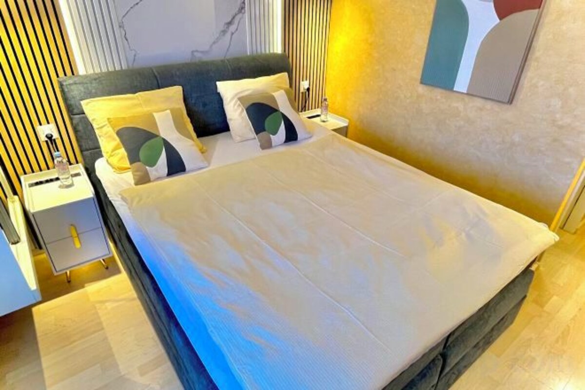 A neatly made bed with a white duvet and pillows, a bedside table with a lamp, and a wall with a painting.
