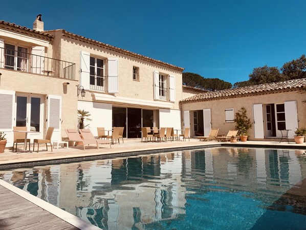 Zona de piscina con sillones reclinables y una mesa de comedor, rodeada por un edificio con tejado de azulejos y contraventanas blancas.