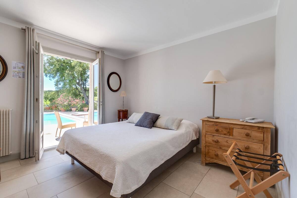 Une chambre avec un lit, une commode en bois, une lampe et une vue sur une piscine à l’extérieur.