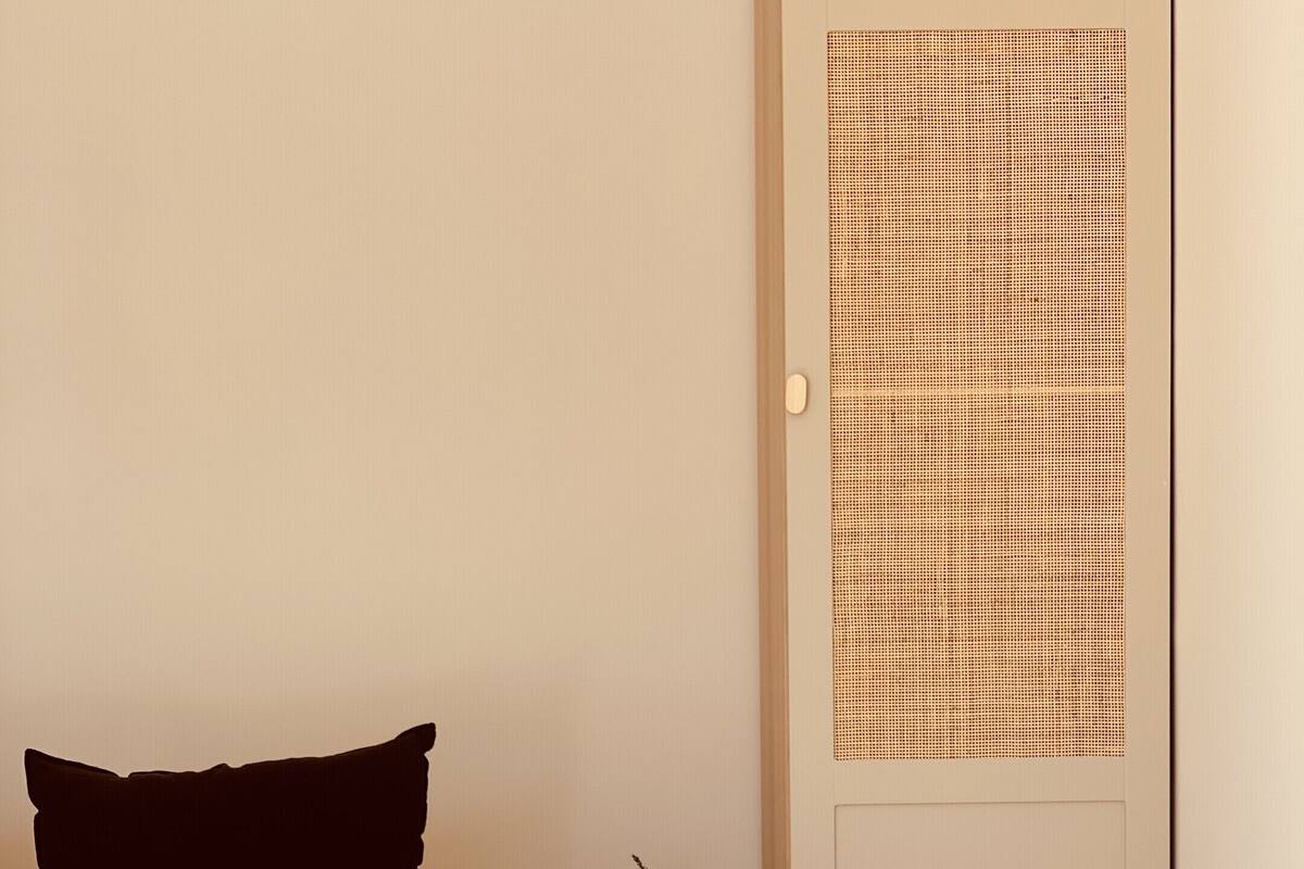 Un lit bien fait, recouvert d’une courtepointe blanche et agrémenté d’un coussin noir, une petite table de chevet avec un vase, et une porte à motifs à carreaux.
