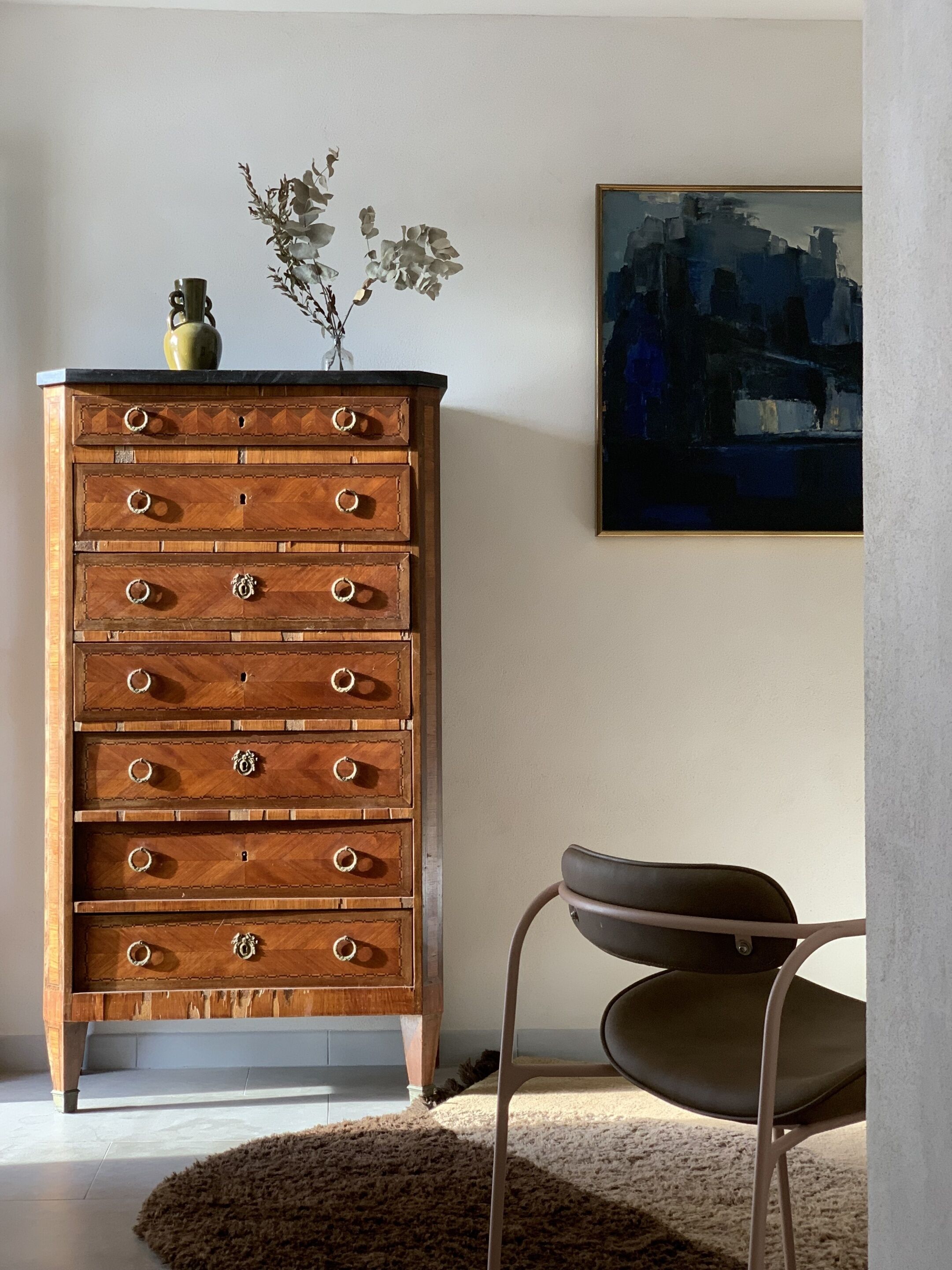 A wooden dresser with multiple drawers, a vase with flowers, and a chair in a room with a painting on the wall.