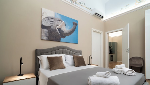 A bedroom with a bed, pillows, a nightstand, a lamp, and a painting of an elephant on the wall. - Archimedes suites  (Catania)