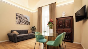 A modern living room with a glass-top dining table, teal chairs, a gray sofa, and a flat-screen TV mounted on the wall. - Archimedes suites  (Catania)