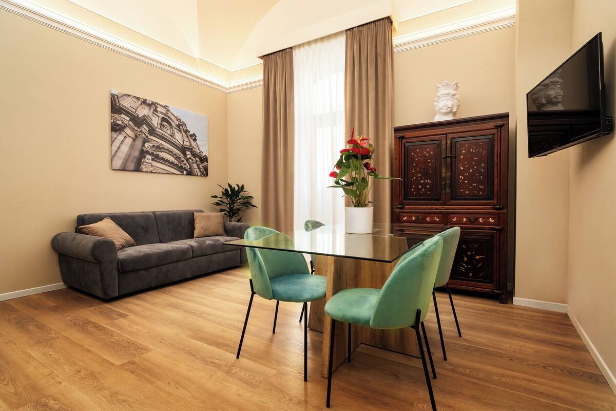 A modern living room with a glass-top dining table, teal chairs, a gray sofa, and a flat-screen TV mounted on the wall.