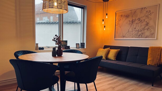 A modern living room with a round wooden table, black chairs, a dark sofa with yellow cushions, a hanging woven light fixture, and a framed abstract artwork on the wall. - Cozy Flats Silkeborg - S3 (Silkeborg)