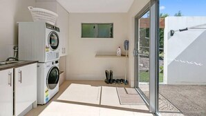 A compact laundry room with a washing machine, a dryer, and a drying rack. There is a small shelf with a bottle and a pair of boots. A window provides a view of the outdoors. - Cooinda Luxury Residence and Studio (Valdora)