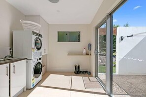 Ein kleiner Waschraum mit Waschmaschine, Trockner und Wäscheständer. Eine kleine Ablagefläche zeigt eine Flasche und ein Paar Stiefel. Ein Fenster bietet einen Blick ins Freie.