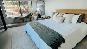 A modern bedroom with a large bed, a bedside table, a mirror, and a view of the outdoors through a large window.