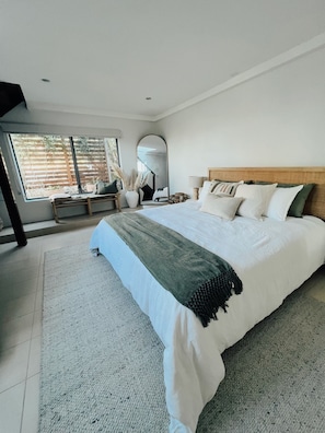 A modern bedroom with a large bed, a bedside table, a mirror, and a view of the outdoors through a large window.