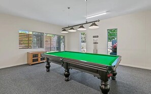Een pooltafel met hangende verlichting, een kast en ramen met jaloezieën.