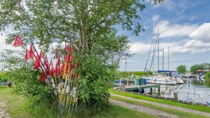 A riverside area with boats docked, a tree with red flags, and a clear sky. - Vacation home Nordhorn - your cozy home in Koserow on the Baltic Sea (Ostseebad Kölpinsee)