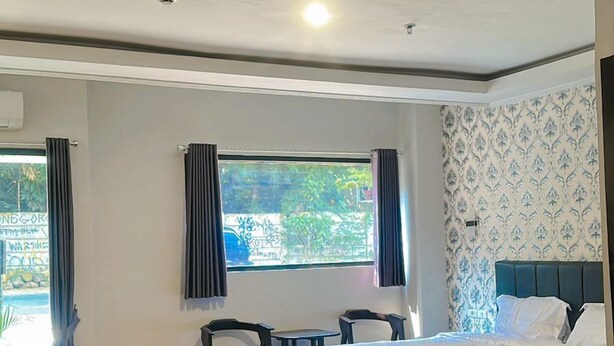 A modern hotel room with a large bed, a small table, and a chair. There is a window with curtains, and the room has a tiled floor.