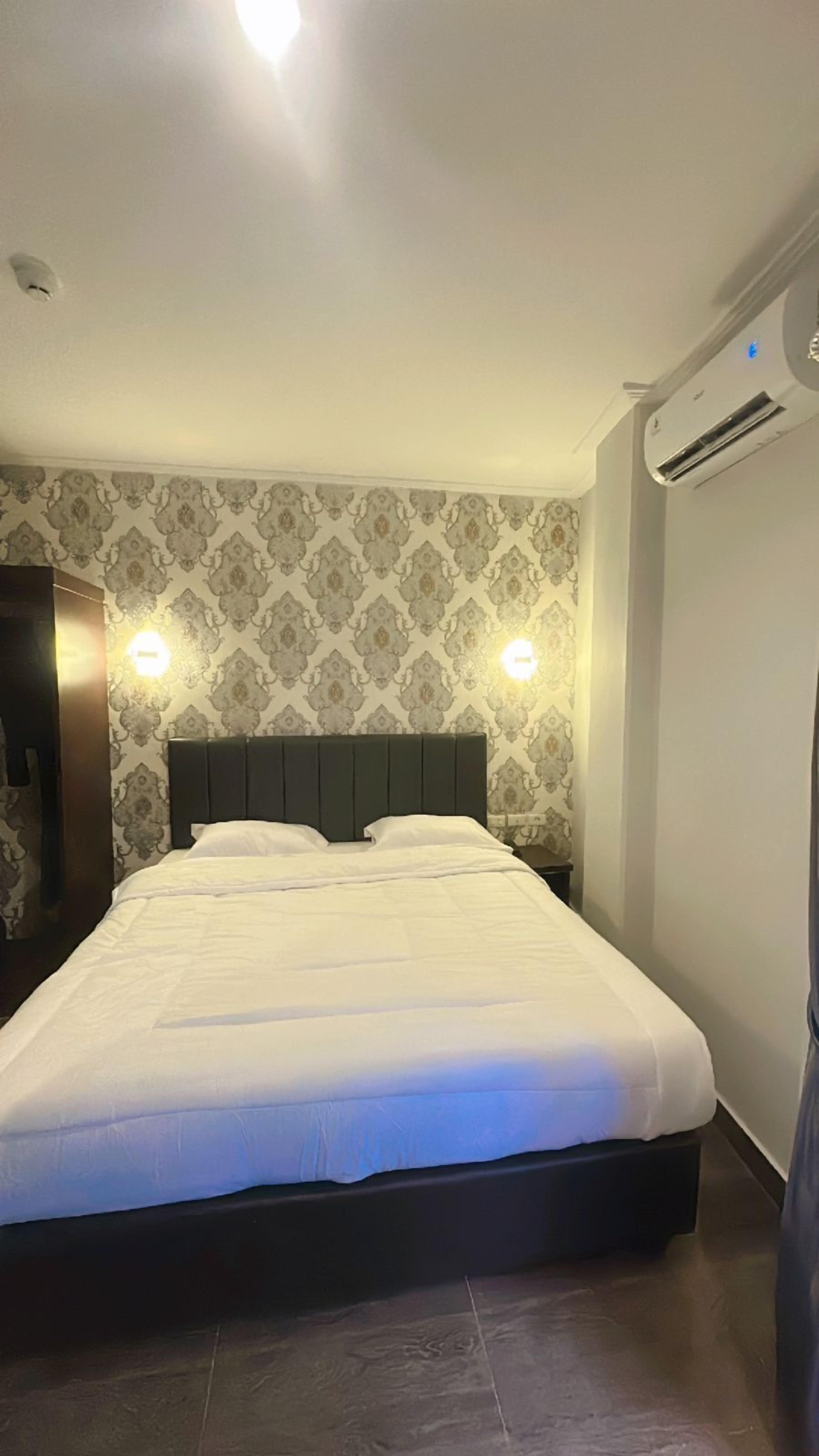 A neatly made bed with a dark headboard in a room with patterned wallpaper and an air conditioning unit on the wall.