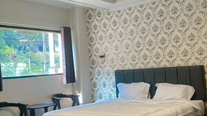 A hotel room with a bed, a small table, and a chair. There is a window with curtains, and the walls have a patterned wallpaper.