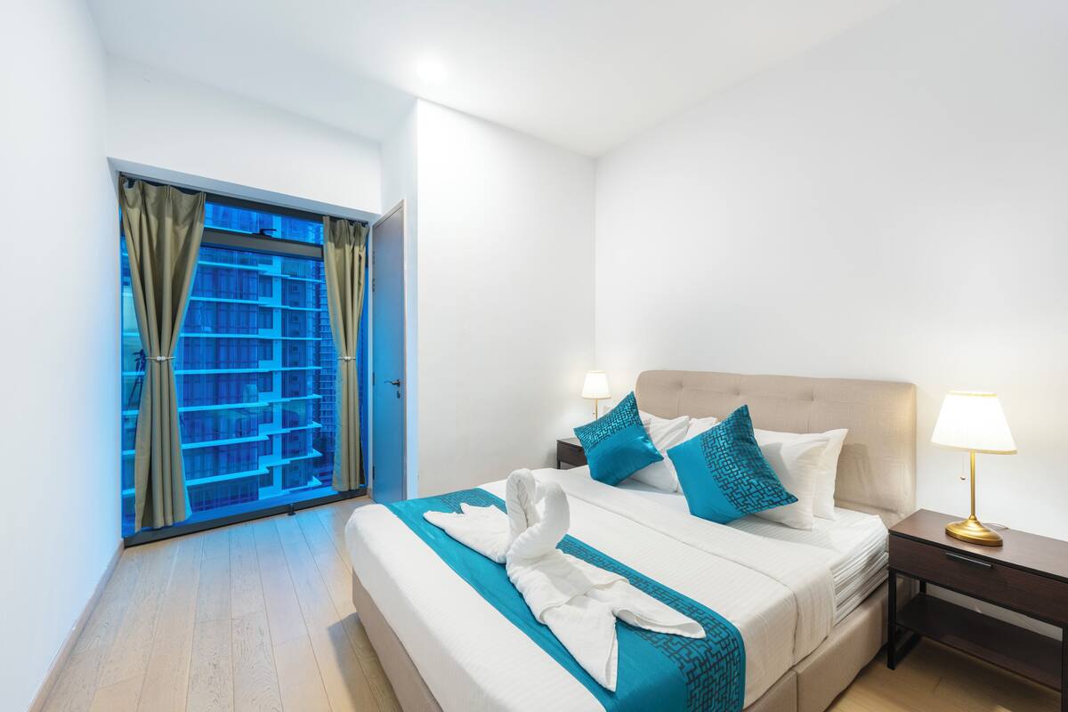 Habitación de hotel con cama, mesita de noche, lámpara, ventana con cortinas y vista a un edificio.