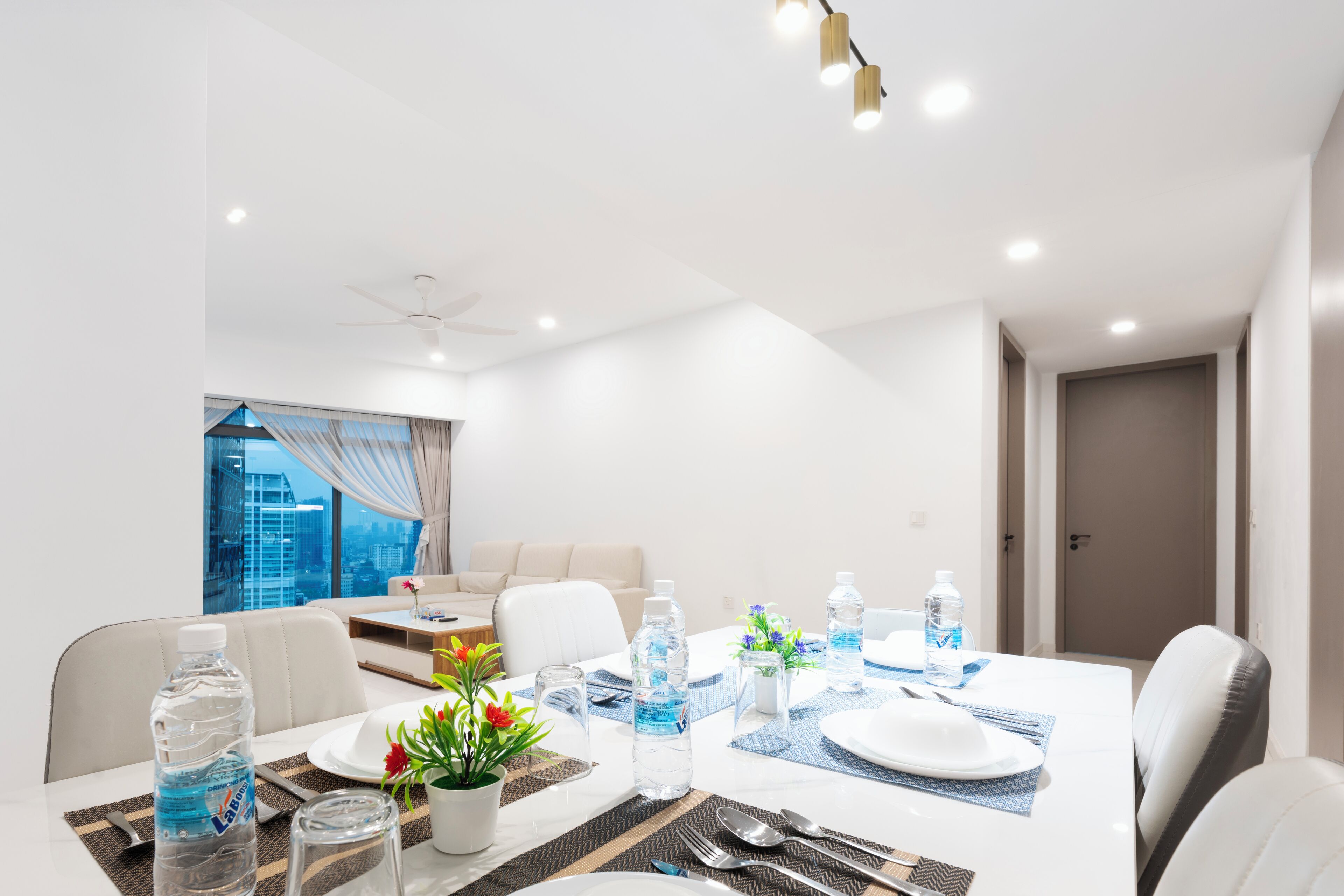 Comedor moderno de hotel con una mesa blanca para cuatro, botellas de agua, una planta en maceta y vista a la ciudad a través de amplias ventanas.