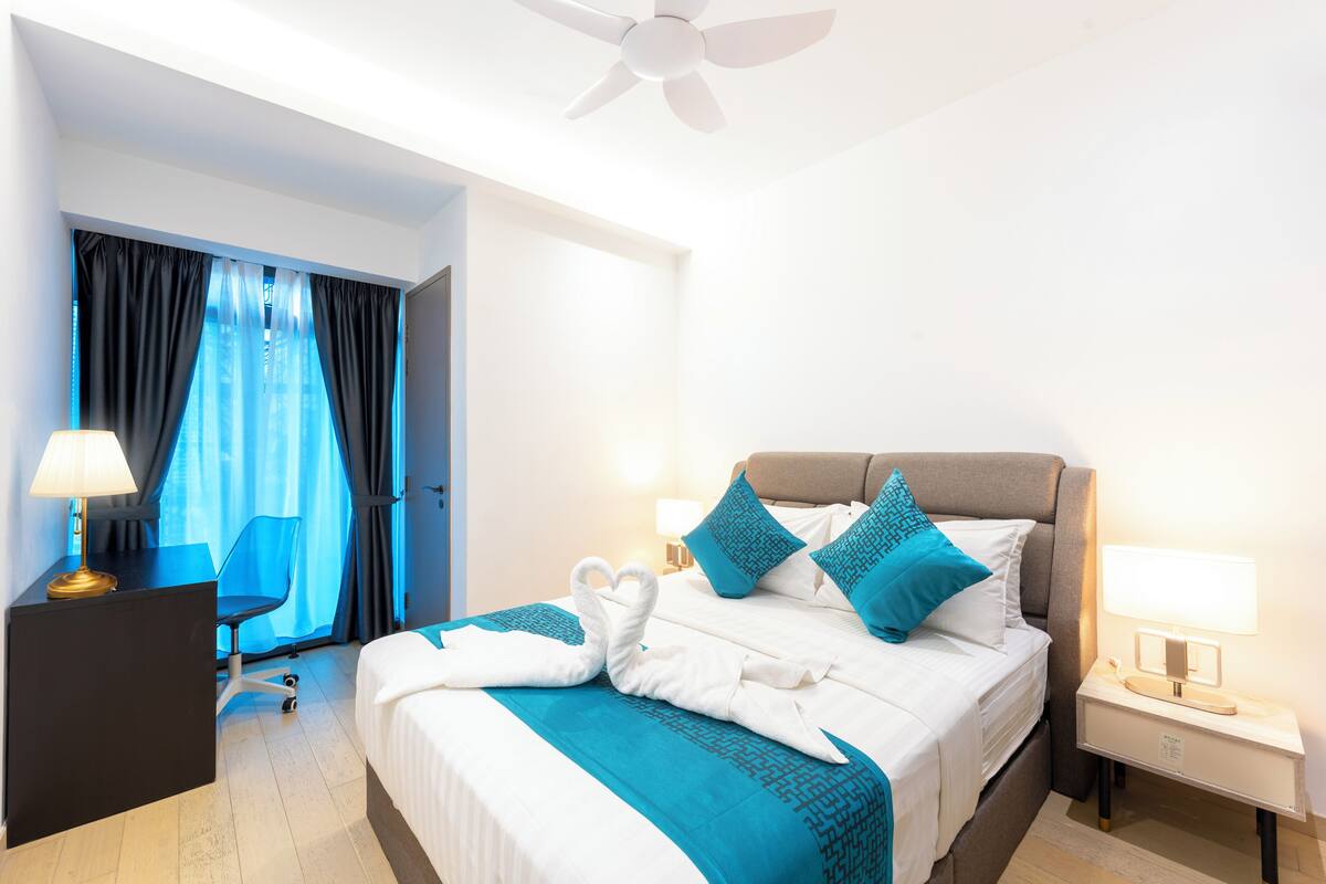 Habitación de hotel con cama, escritorio, silla, lámpara de escritorio, mesita de noche con lámpara y ventilador de techo.