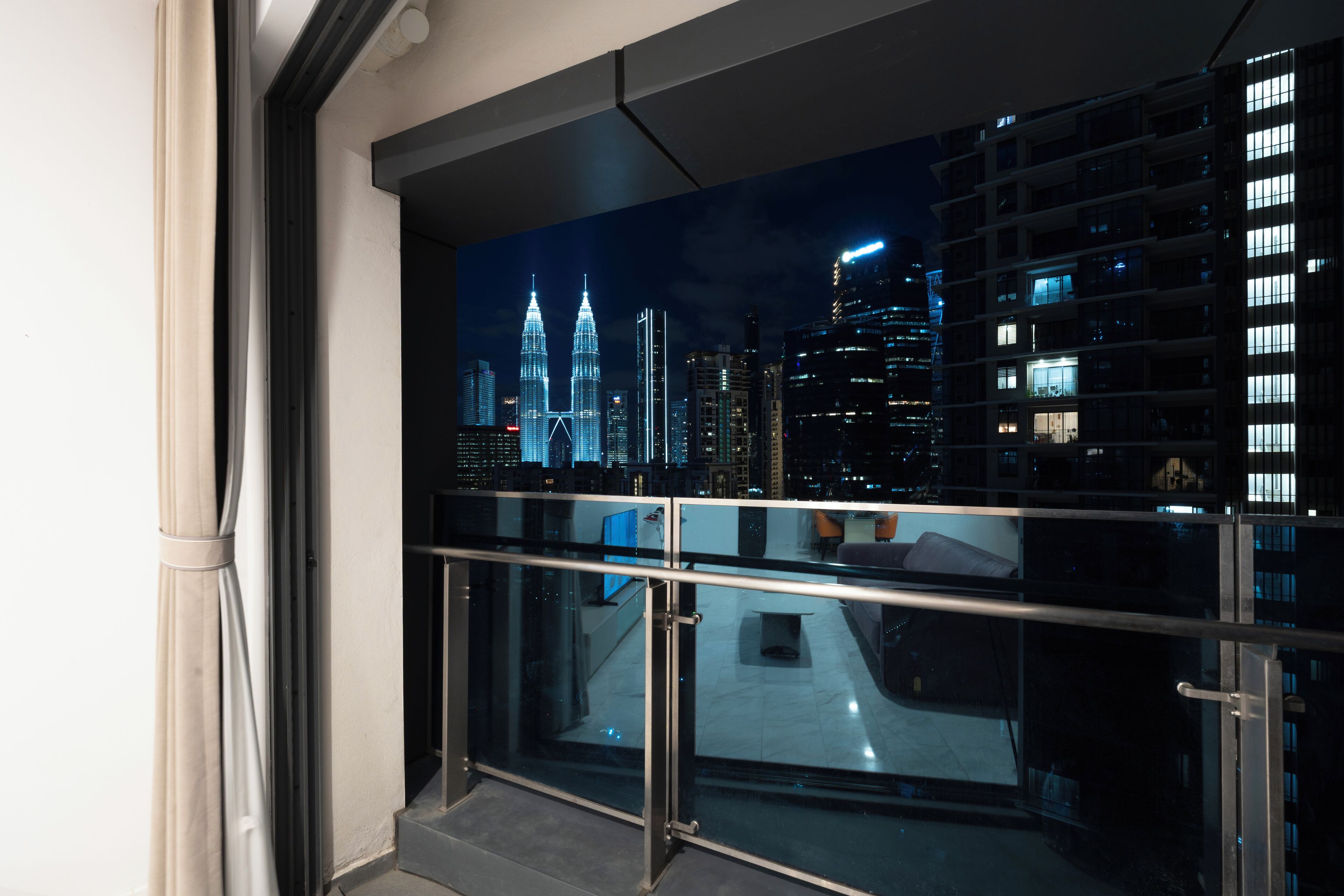 A view from a balcony with a glass railing, overlooking a cityscape at night with illuminated buildings.