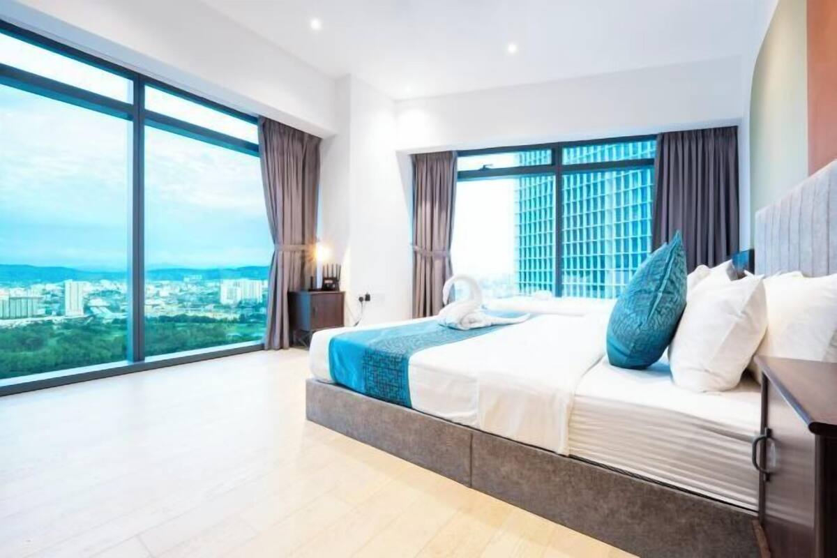 Una habitación de hotel moderna con una cama grande, una mesita de noche, una lámpara y vistas a la ciudad a través de amplios ventanales.
