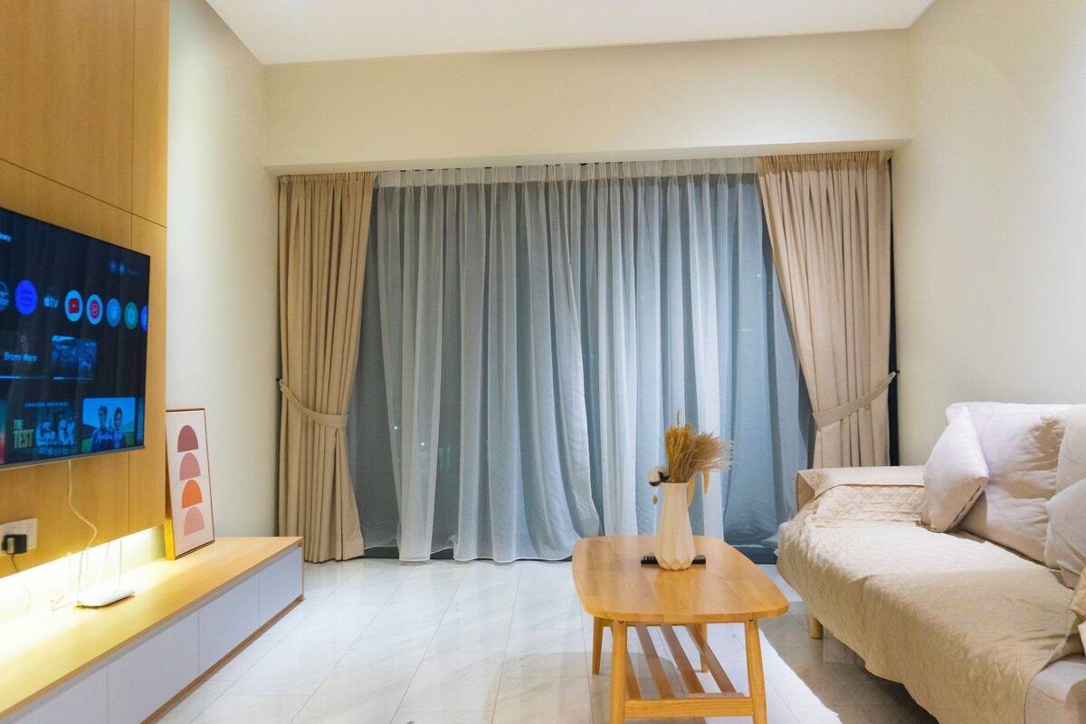 Una sala de estar moderna con un televisor de pantalla plana, un sofá, una mesa de centro y un amplio ventanal con cortinas.