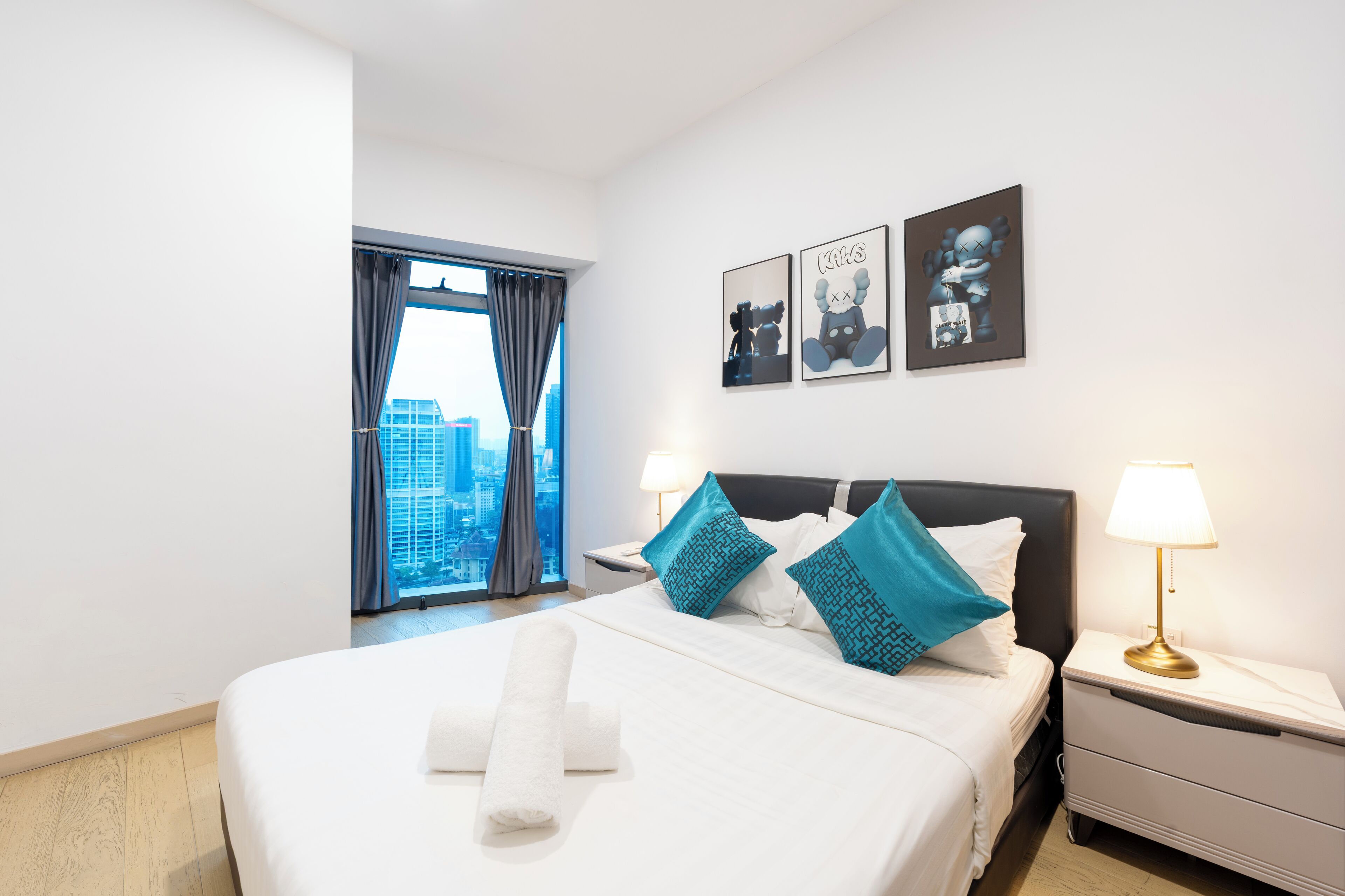 A hotel room with a large bed, two pillows, a nightstand with a lamp, a window with curtains, and two wall art pieces.