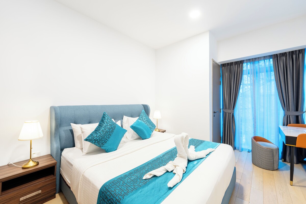 Una habitación de hotel con cama, escritorio, silla, lámpara y ventana con cortinas.