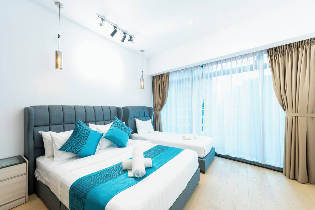 Habitación de hotel con dos camas, un ventanal grande con cortinas y un moderno aplique de techo.