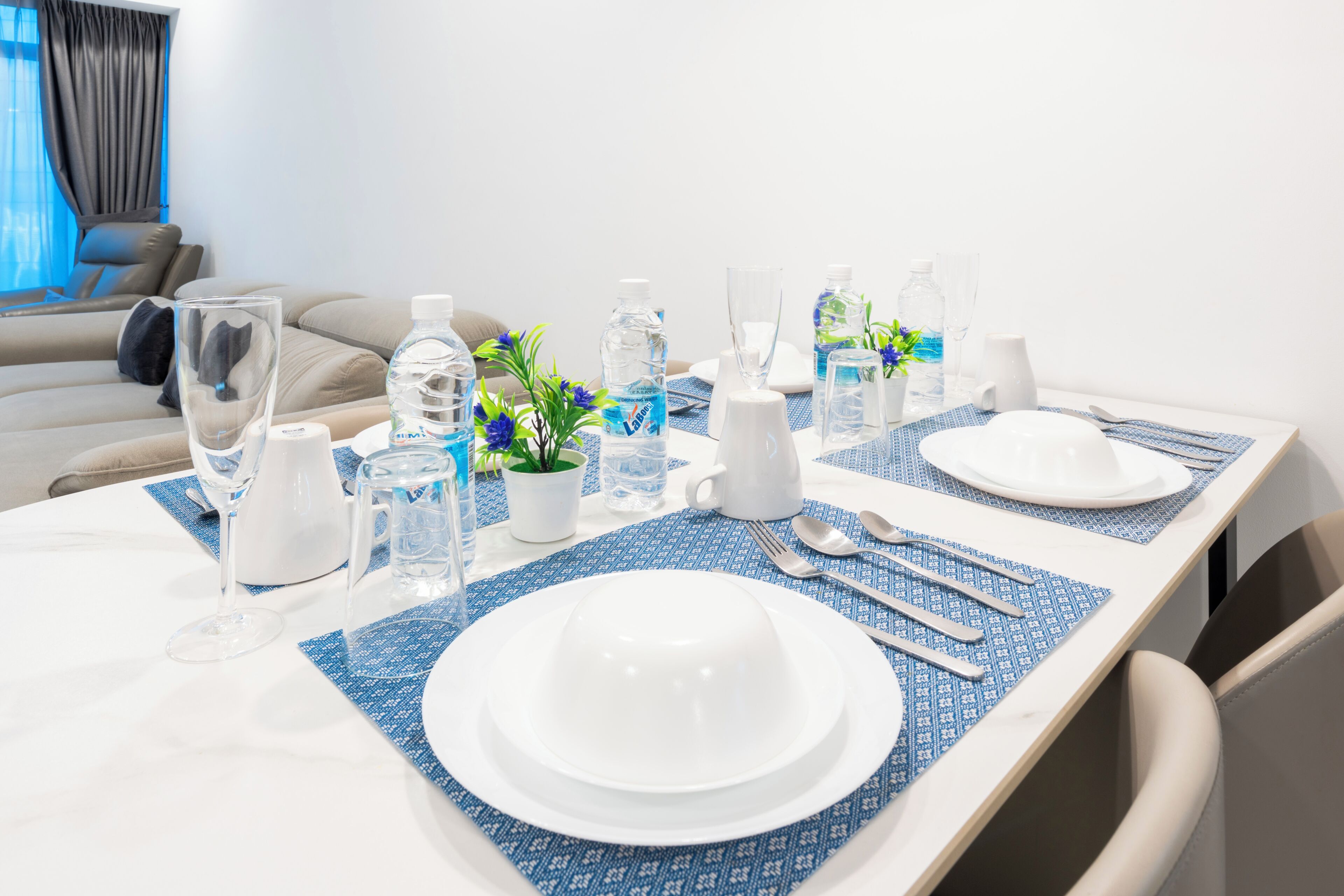 Una mesa de comedor dispuesta con platos blancos, cubiertos y botellas de agua, con vista a una sala de estar al fondo.
