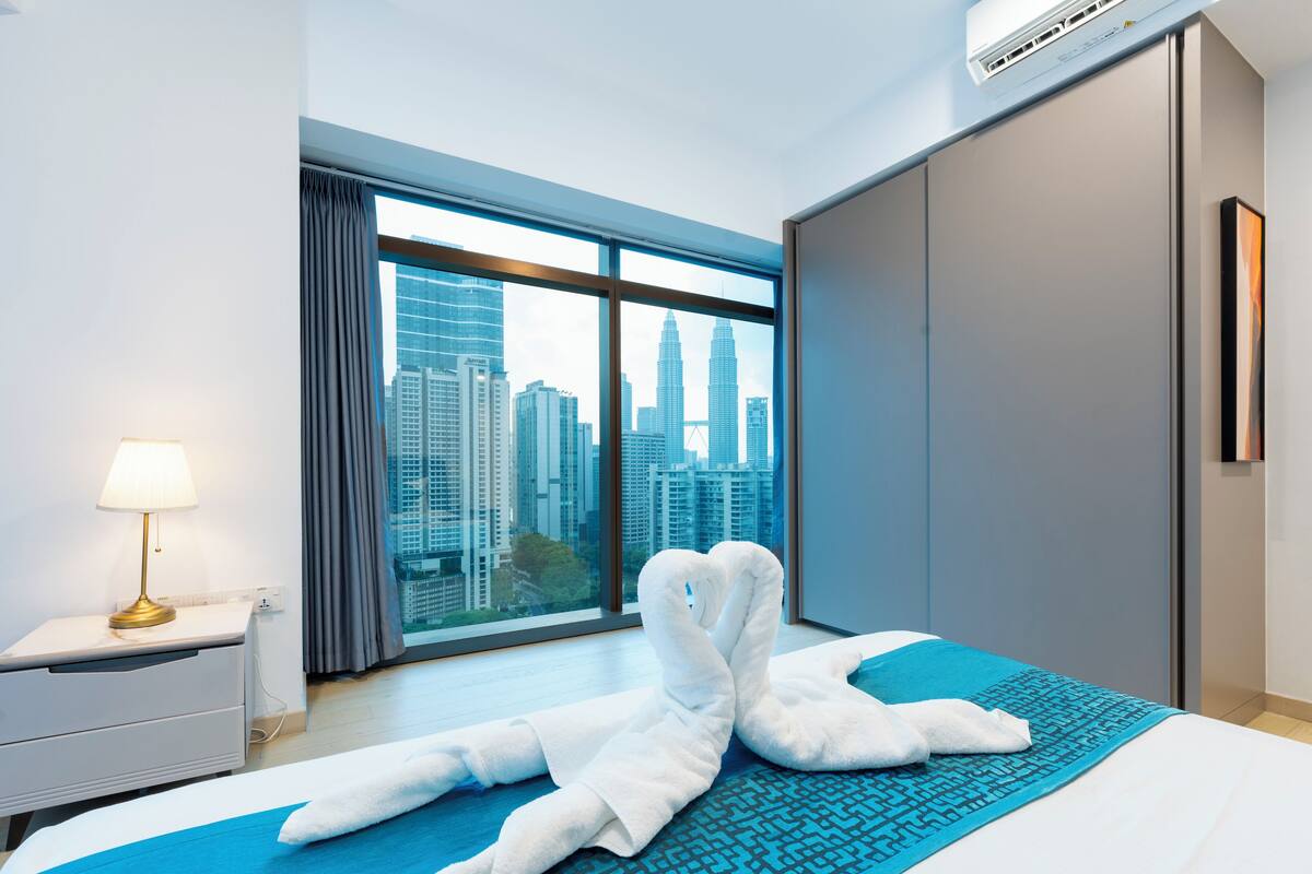 Habitación de hotel con cama, mesita de noche, lámpara, ventana con cortinas y vista panorámica de la ciudad.