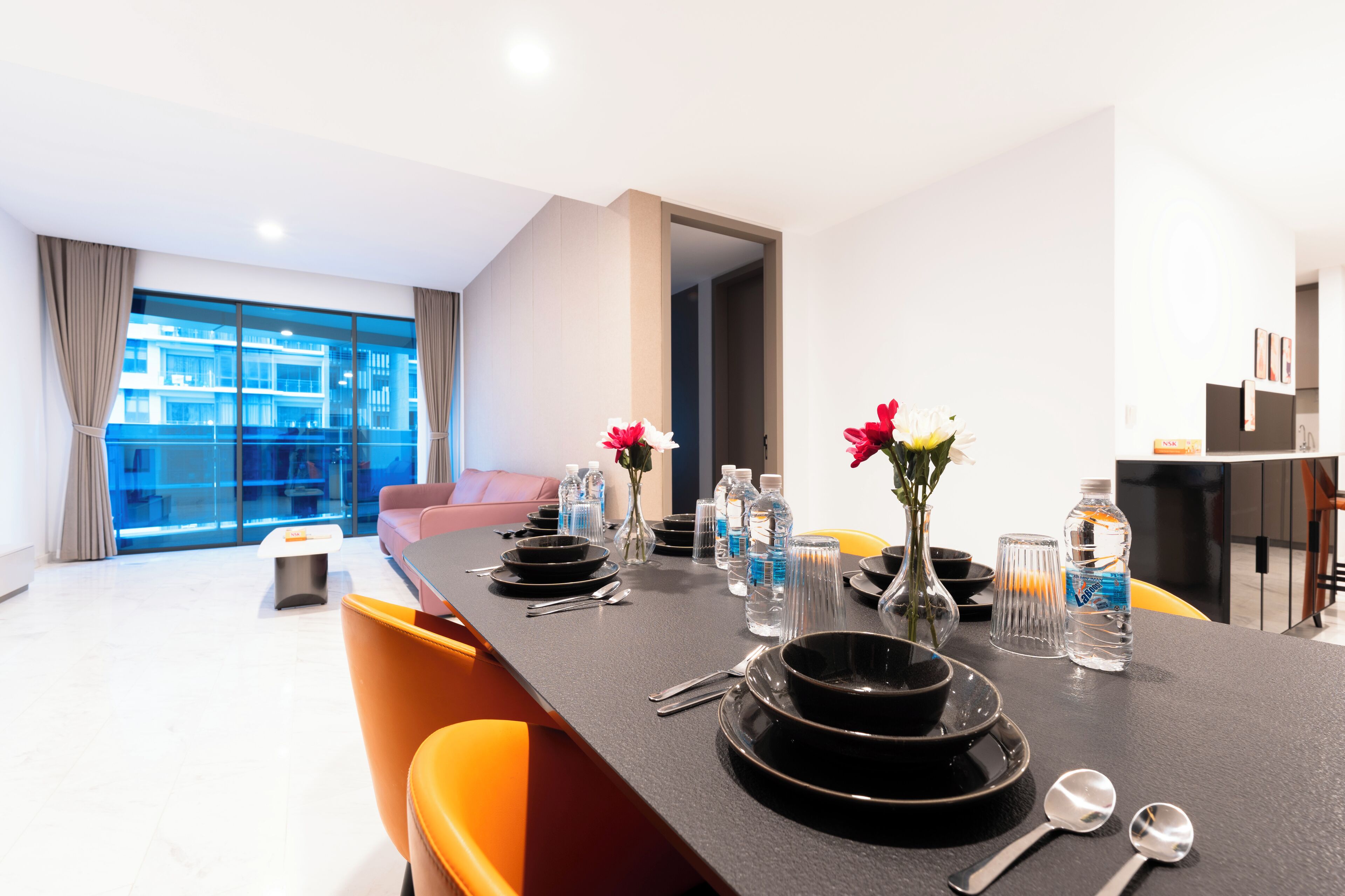 A modern dining area with a long table set for a meal, featuring clear glassware, black plates, and a vase with flowers.