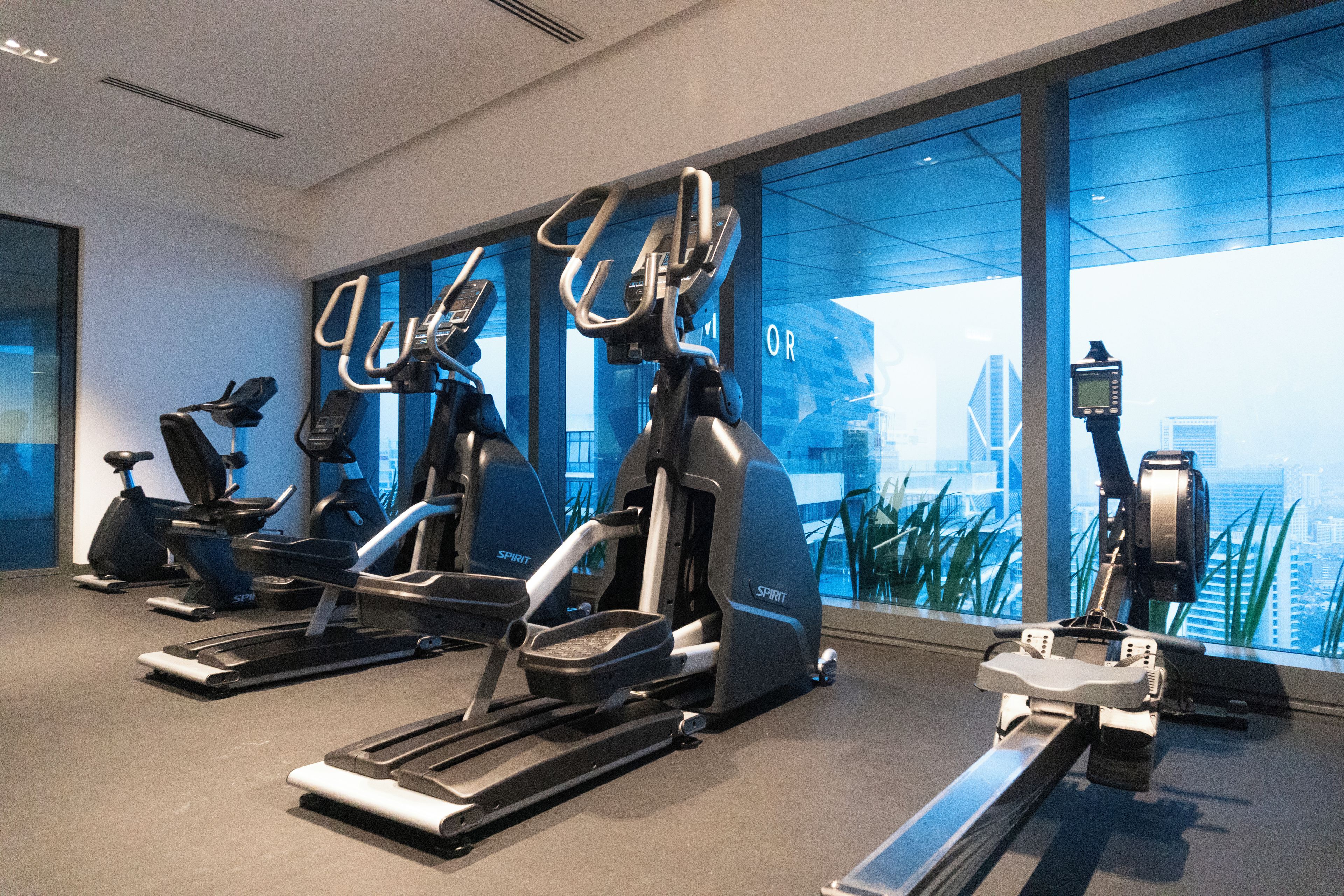 Un gimnasio con diversos equipos de ejercicio, incluyendo cintas de correr y bicicletas estáticas, amplias ventanas con vista a la ciudad y un diseño interior moderno.