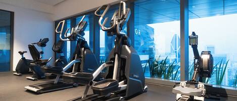Un gimnasio con diversos equipos de ejercicio, incluyendo cintas de correr y bicicletas estáticas, amplias ventanas con vista a la ciudad y un diseño interior moderno.