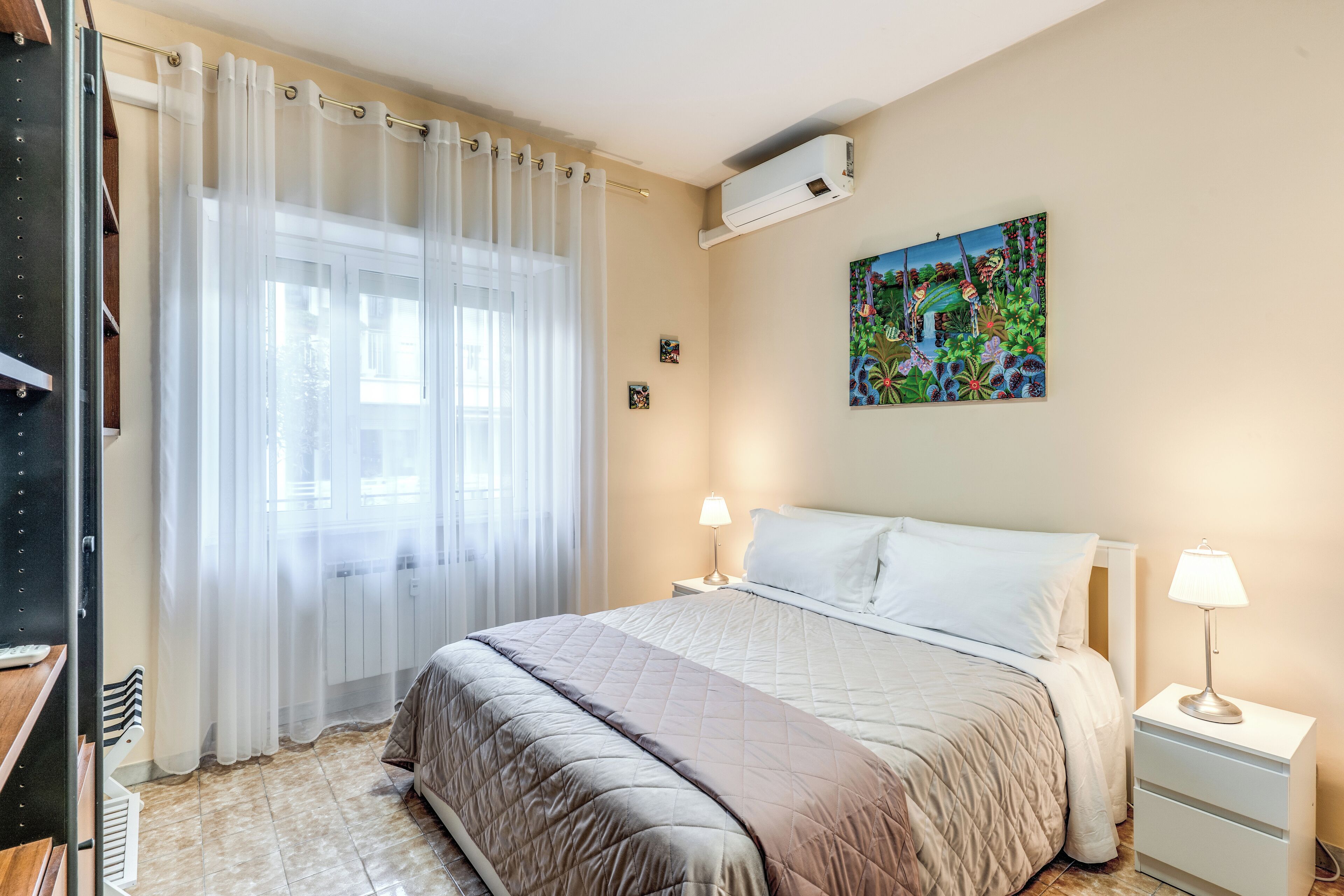 A bedroom with a bed, bedside tables, a window with curtains, a wall-mounted air conditioner, and a painting.