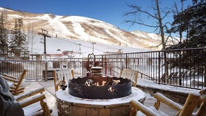 Un espacio al aire libre cubierto de nieve con un hogar, sillas de madera y vistas a una pista de esquí al fondo.